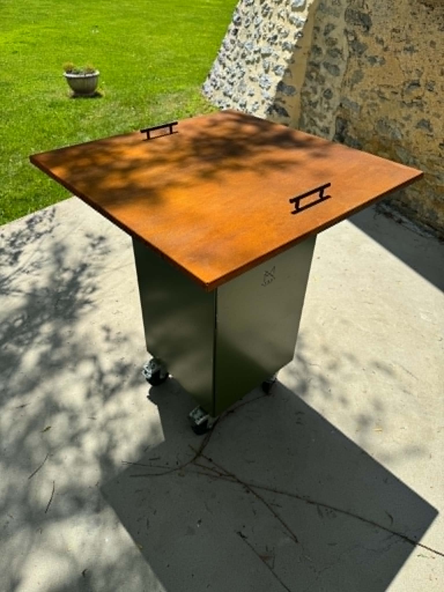 Rust-colored metal outdoor table with a pedestal base and small wheels, set on a sunny concrete patio by a stone wall and green lawn.