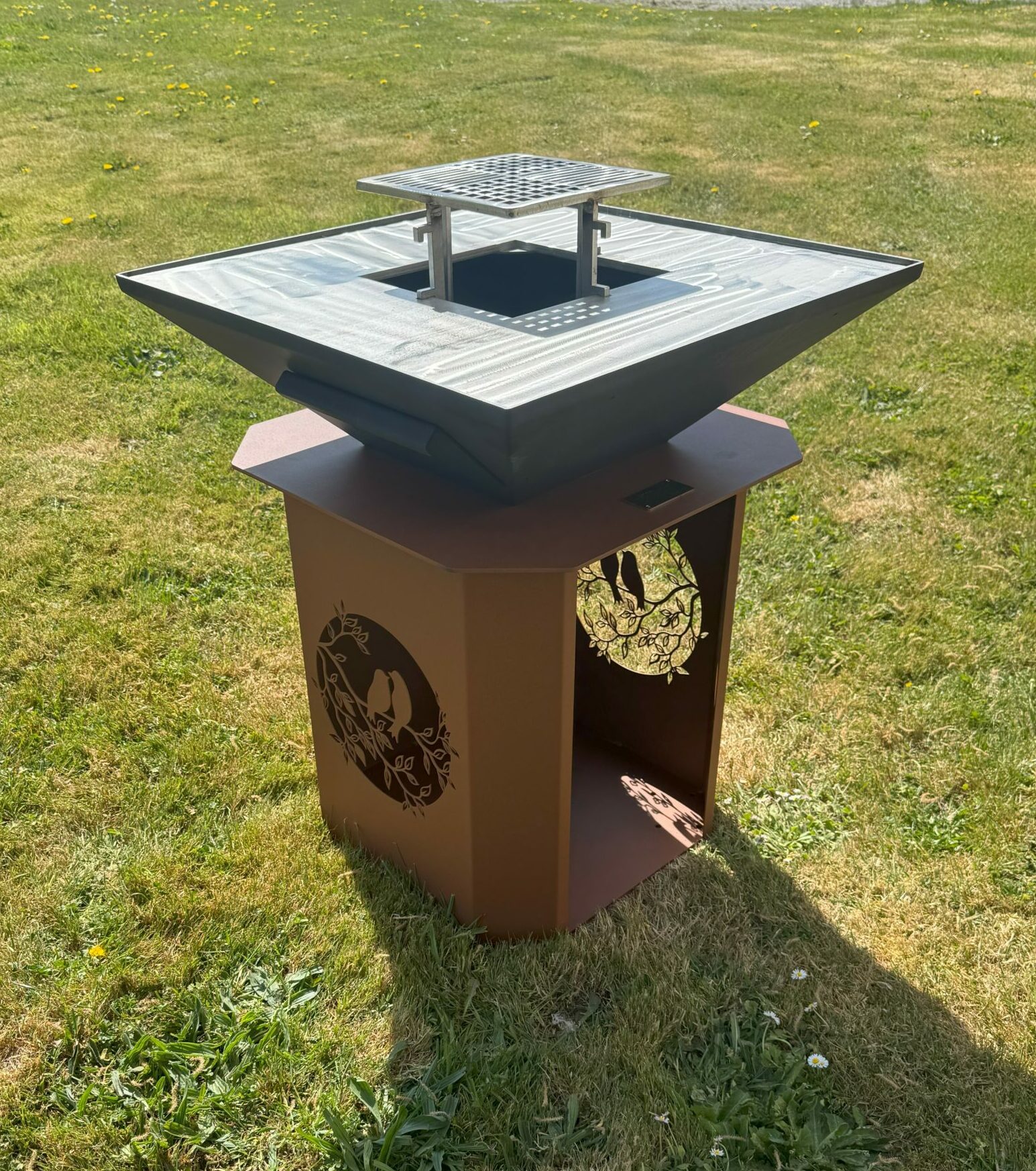 Outdoor metal sculpture in a grassy park: a square grate atop a tilted, pyramid-like metal body mounted on a brown pedestal.