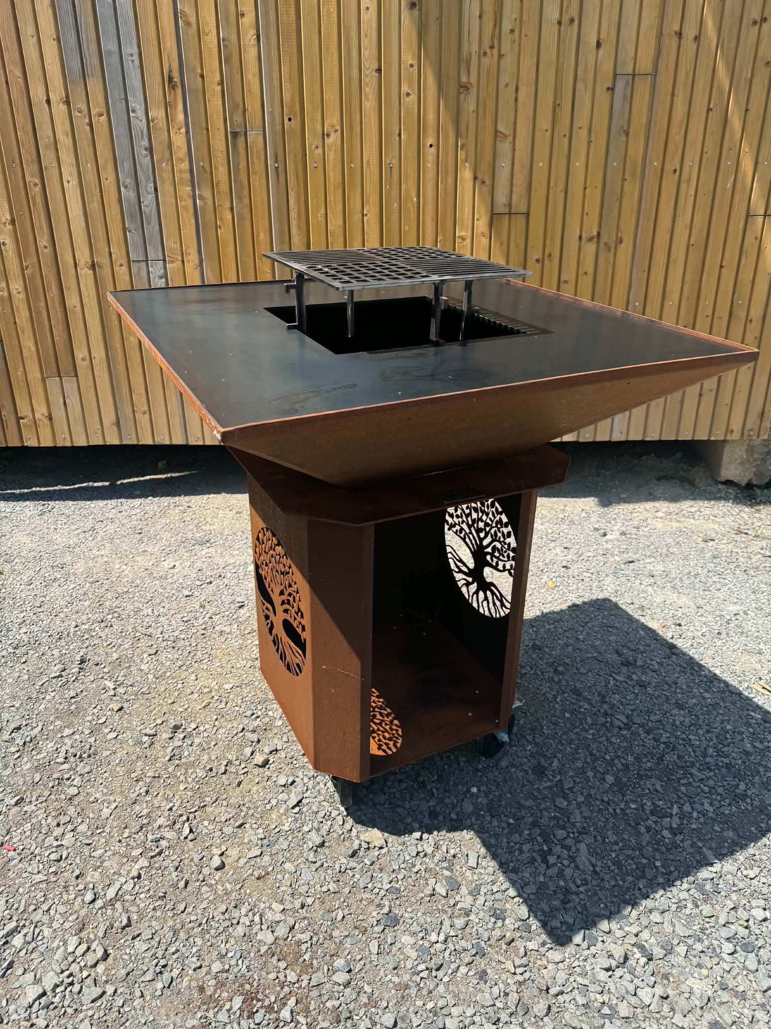Rusty metal fire pit with a square grate and tree-cutout panels, set on gravel in front of a wooden fence.